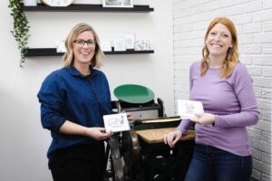 Hannah and Amanda hold their custom stationary in front of the letterpress machine Cornerstone Press + Studio A Branding Session