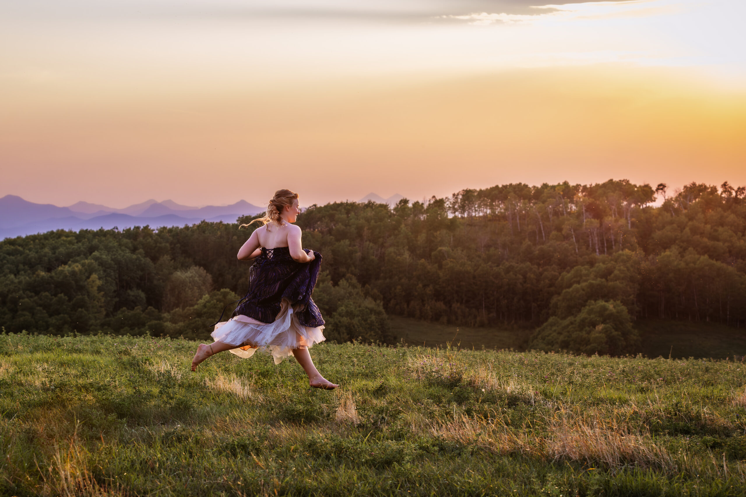 calgary high school grad running through field in grad dress with rocky mountains in background