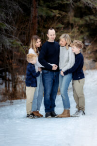 A family of five embraces tightly on a snow-covered forest path, surrounded by tall evergreen trees. Their warm smiles and cozy winter outfits create a feeling of love and togetherness. The soft light and untouched snow add a serene and magical atmosphere to the scene.