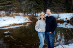A man and woman stand close together outdoors, smiling gently at the camera. The woman is dressed in a white knit sweater and jeans, while the man wears a black sweater and jeans. Behind them, a calm creek reflects the surrounding snow-covered forest.