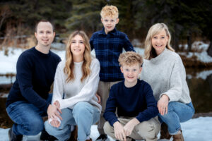 A family of five crouches together in a classic pose near a calm, reflective pond. They are close to one another, with warm smiles and coordinated winter outfits. The snowy ground and tall evergreens in the background frame the scene beautifully, creating a timeless and serene family portrait in Griffith Woods Calgary Alberta