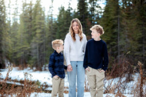 Three siblings stand together on a snowy riverbank, sharing a moment of laughter. The water sparkles behind them, reflecting the soft golden tones of the late afternoon light.