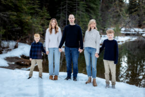 A family of five stands together holding hands near a serene pond, all looking at the camera with warm smiles. The snow-covered ground and evergreen trees reflect the soft winter light, creating a peaceful and inviting scene.