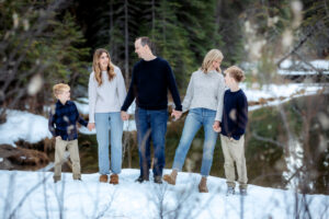 A family of five holds hands near a tranquil pond, laughing and smiling at one another. Their joyful connection shines through as they share a lighthearted moment, surrounded by snowy ground and the natural beauty of the forest.