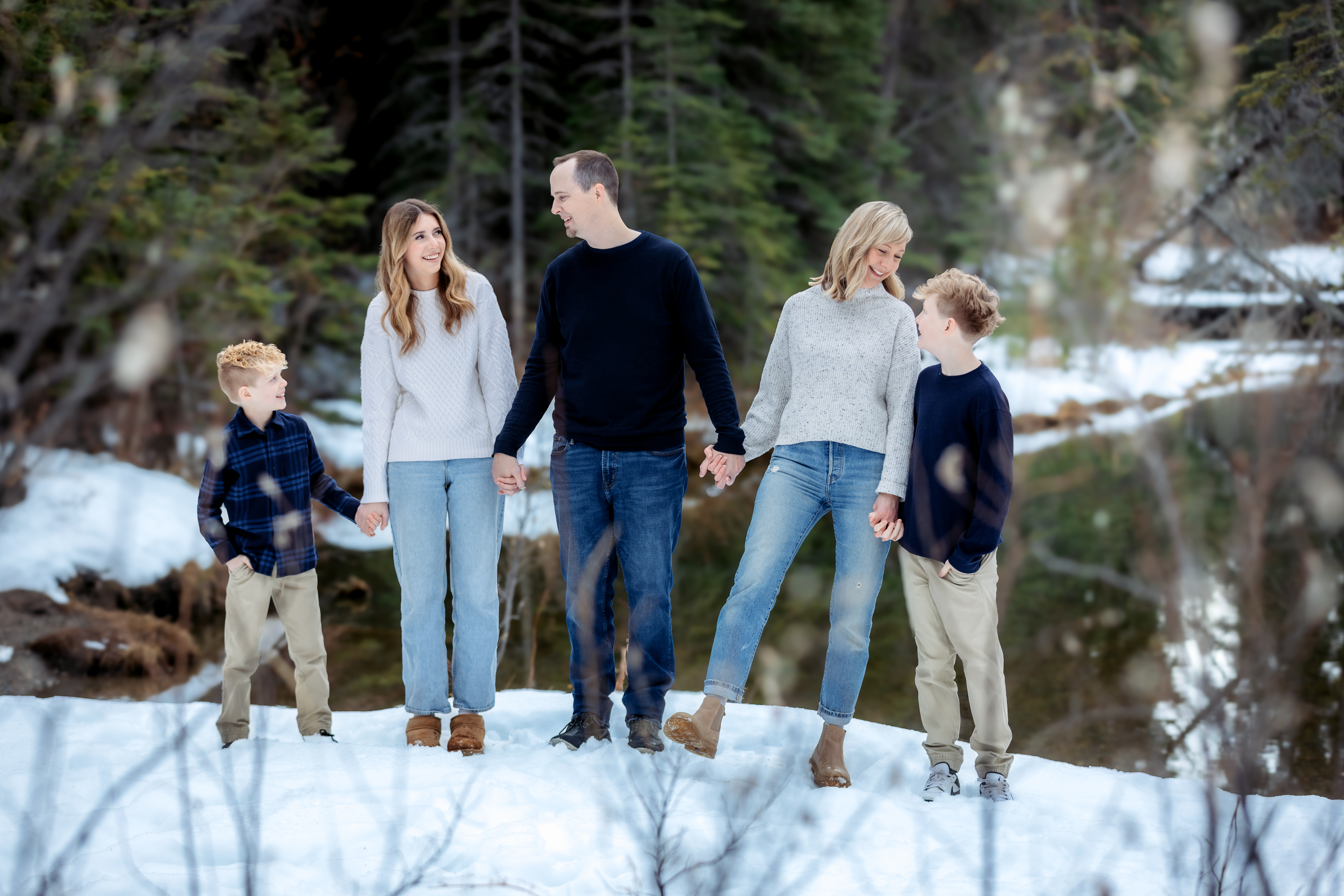 A family of five holds hands near a tranquil pond, laughing and smiling at one another. Their joyful connection shines through as they share a lighthearted moment, surrounded by snowy ground and the natural beauty of the forest.