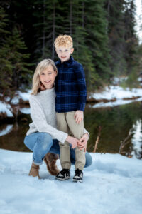 A woman kneels in the snow next to her son, her arm resting around him lovingly. She wears a gray sweater, jeans, and brown boots, while the boy is dressed in a blue plaid shirt and khaki pants. They are surrounded by a serene winter landscape of trees and a still river.