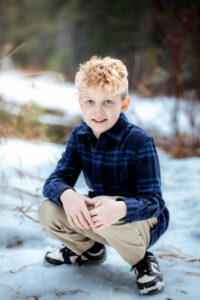 A young boy with curly blonde hair crouches on a snowy path, wearing a blue plaid shirt, khaki pants, and sneakers. His bright blue eyes and warm smile stand out against the snowy forest backdrop. The scene is softly lit, with blurred greenery in the background.