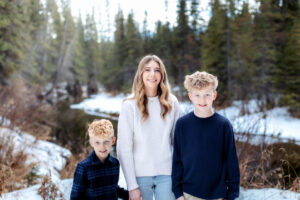 Three siblings stand together on a snowy riverbank, smiling at the camera. The water sparkles behind them, reflecting the soft golden tones of the mid morning light.