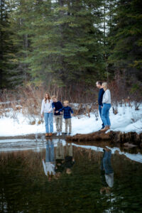 A couple stands side by side, smiling as they watch their three children throwing rocks into a calm pond. The kids, spaced apart along the snowy bank, are mid-motion, with splashes rippling across the water. The surrounding forest and soft winter light create a peaceful, natural setting.