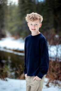 A young boy with curly blonde hair stands on a snowy path by a river bank, wearing a blue sweater, khaki pants, and sneakers. His bright blue eyes and warm smile stand out against the snowy forest backdrop. The scene is softly lit, with blurred greenery in the background.