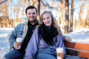 Sitting on a park bench with steaming hot chocolate cups, the couple smiles warmly at the camera. Their casual winter outfits and the serene setting create a cozy, inviting moment.