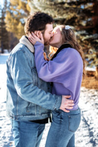 On a snowy path beside the skating pond, the couple shares a tender kiss. She rests her engagement-ring-adorned hand on his neck, and he places his hand on her waist. The sunlight filters through the trees, adding a golden glow to the romantic scene.
