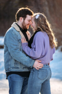 The couple embraces on the ice, gazing deeply into each other’s eyes. She wears a purple sweater, and he has a sherpa-lined jean jacket. Their jeans and wintery surroundings create a romantic and laid-back moment on the frozen pond.