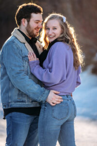 A couple stands on ice skates in a warm embrace. He looks lovingly at her while she gazes at the camera, her purple sweater vibrant against the snowy background. He wears a sherpa-lined jean jacket, and both are in jeans, adding a casual, cozy feel to the scene.