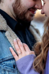 Her hand, adorned with a shimmering engagement ring, rests gently on his chest as they embrace. The close-up captures the warmth of their connection, with his sherpa lined jean jacket providing a textured backdrop to the symbolic ring
