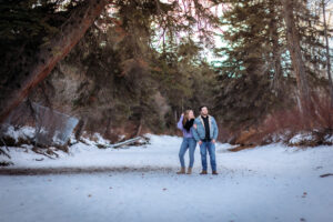 The couple stands on the frozen river under a towering tree, while she is looking away from the photographer. She has one hand in her hair, adding a playful and candid feel to the image. The scene’s vastness highlights their connection amidst the beautiful winter landscape.
