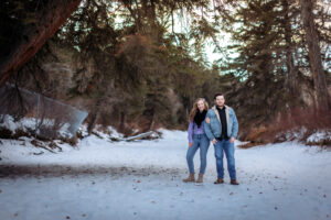 The couple stands close together on a frozen river beneath a massive evergreen tree. The wide angle captures the grandeur of the tree and the surrounding snowy landscape, while the couple’s warm expressions draw focus.