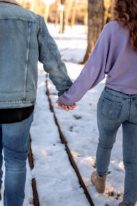 The couple walks hand in hand along a snow-dusted train track surrounded by winter trees. Their steps are in sync, and their backs are to the camera. They convey a sense of togetherness in the serene park environment.