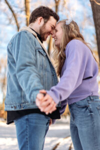 A couple skates on a frozen pond, holding hands extended toward the camera. Their faces are in focus, radiating joy and excitement. The wintery backdrop of trees and soft light enhances the romantic atmosphere.