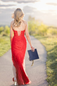 Calgary grad walking along a pathway at sunset in a red dress holding her graduation cap with mountain views in the background